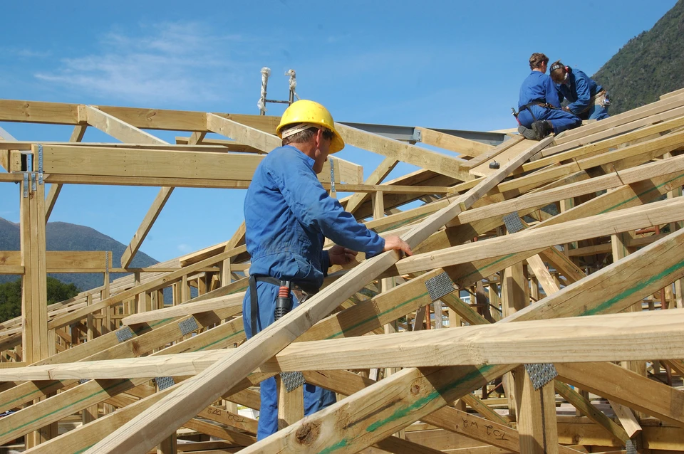 Chantier de construction extérieur avec ouvriers en combinaisons bleues et casques jaunes travaillant sur charpente bois